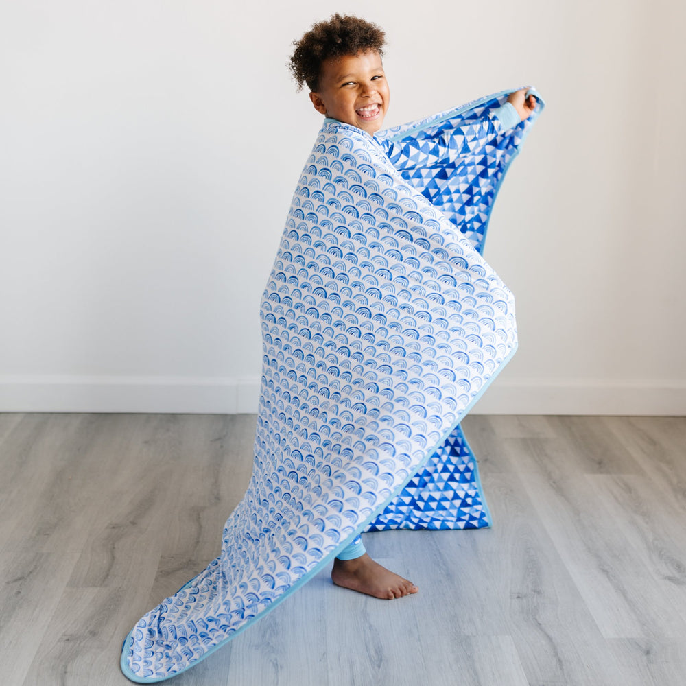 Image of little boy draping the blanket over his shoulders. The image features the blanket being reversible, with one side showing blue rainbows and the other side showing blue horizon triangles. The two prints feature different shades of blue.