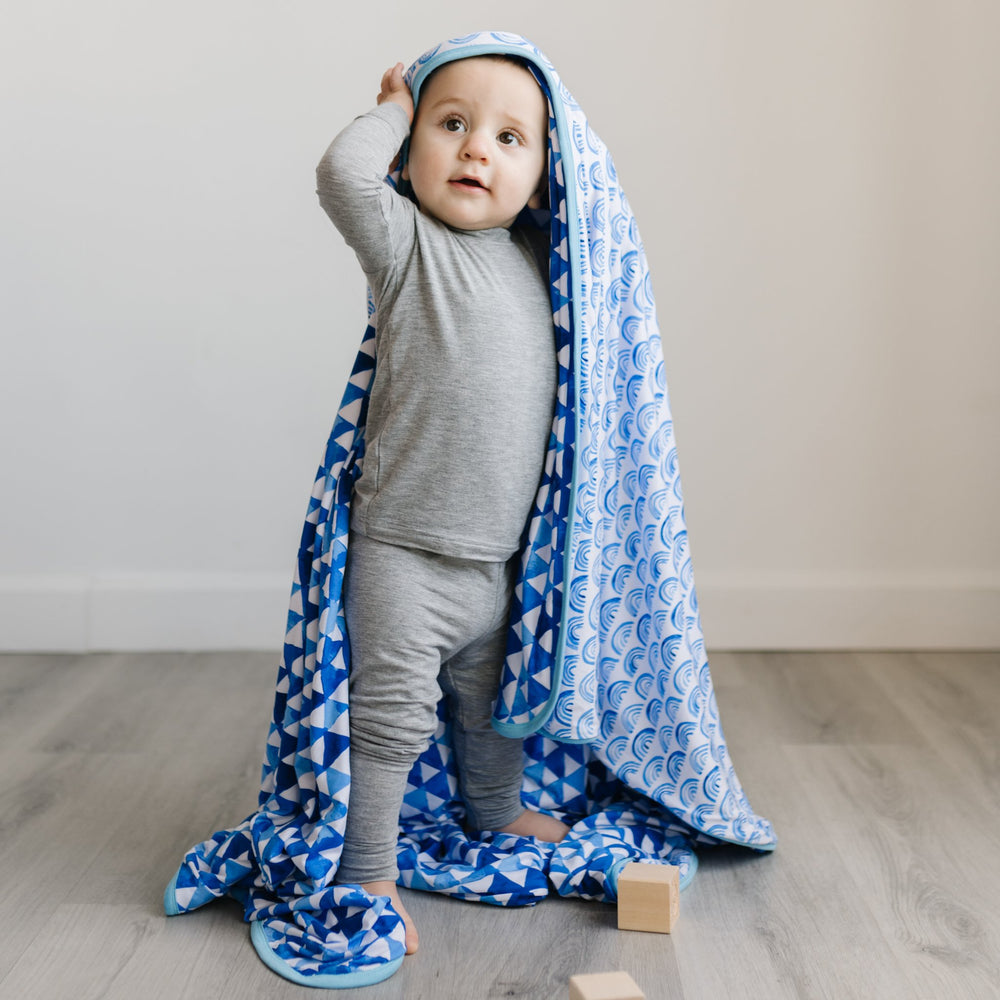 Image of little boy draping the blanket over his head. The image features the blanket being reversible, with one side showing blue rainbows and the other side showing blue horizon triangles. The two prints feature different shades of blue.