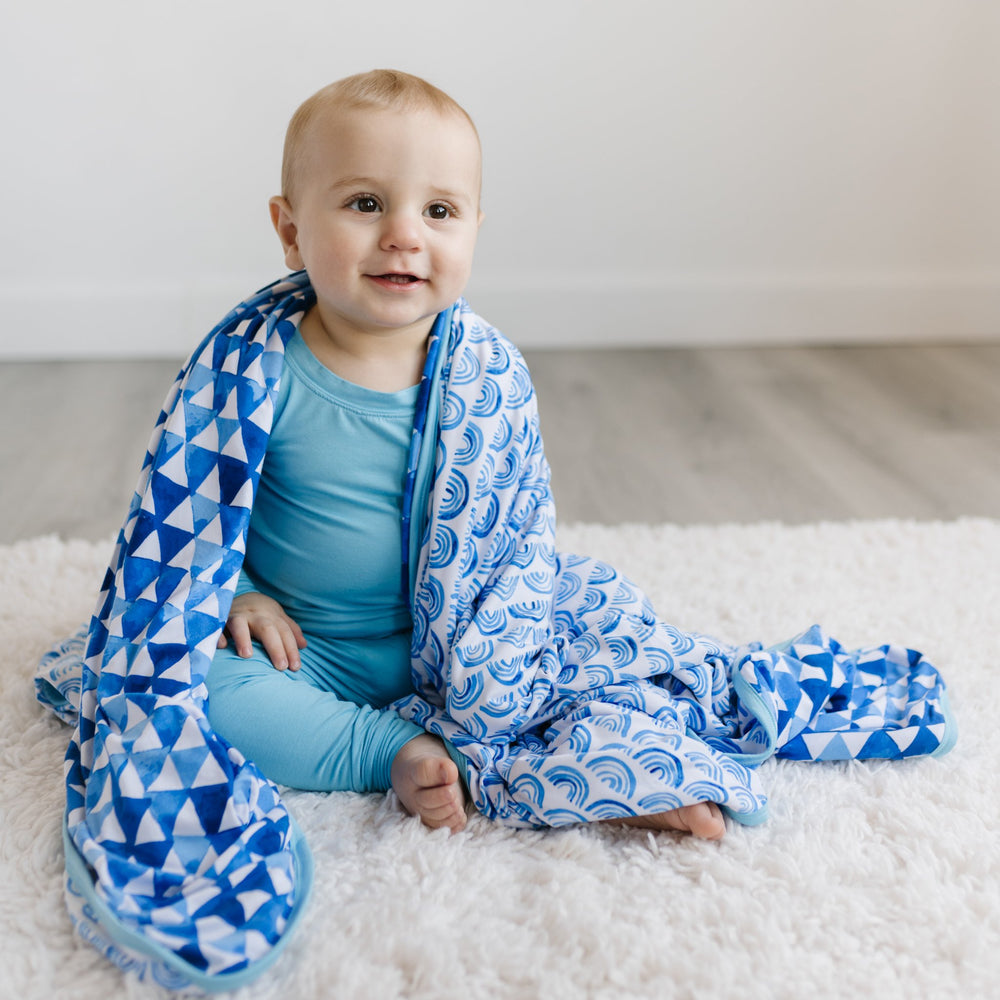Image of little boy with the blanket draped over his shoulders. The image features the blanket being reversible, with one side showing blue rainbows and the other side showing blue horizon triangles. The two prints feature different shades of blue.