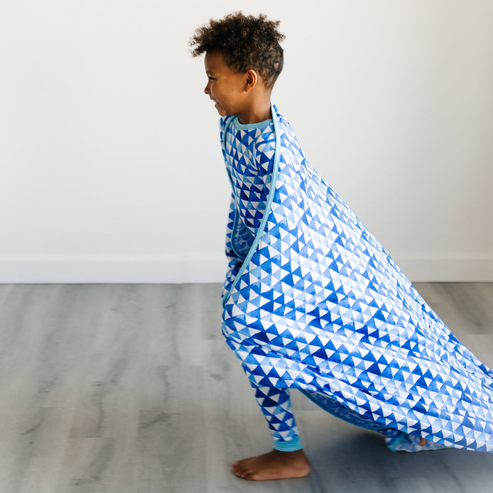 Image of little boy with blanket draped over his shoulders. He is featured wearing blue horizon triangles pajama set, which match the blue horizon triangles print on the blanket. The blanket features different shades of blue. 