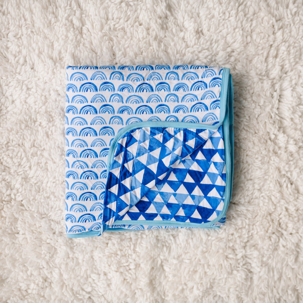 Click to see full screen - Flat lay photo of blanket folded into a square. The image features the blanket being reversible, with one side showing blue rainbows and the other side showing horizon triangles. The two prints feature different shades of blue.