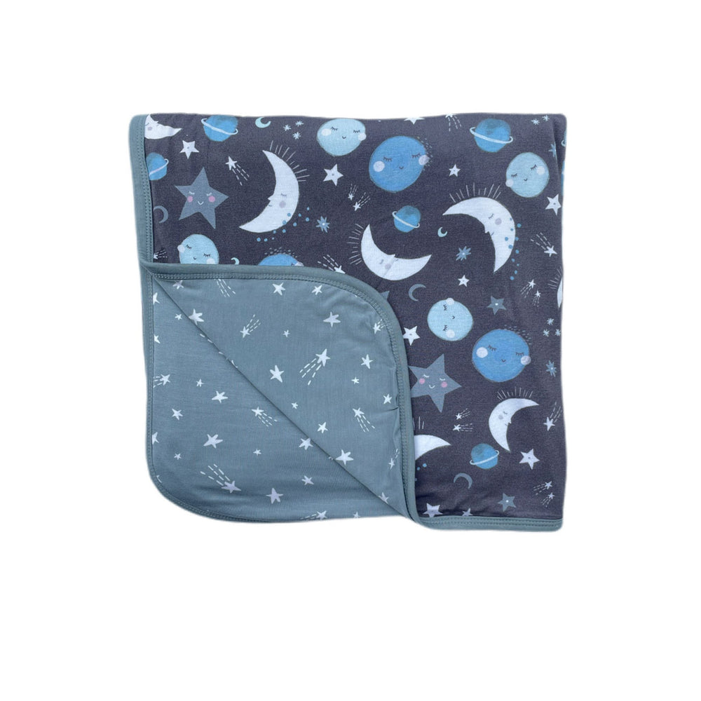 Flat lay image of large cloud blanket in blue to the moon and back print. This blanket features a double-sided design, with blue and gray moons, stars, and planets on a charcoal background on one side and white shooting stars on a coordinating blue backgr