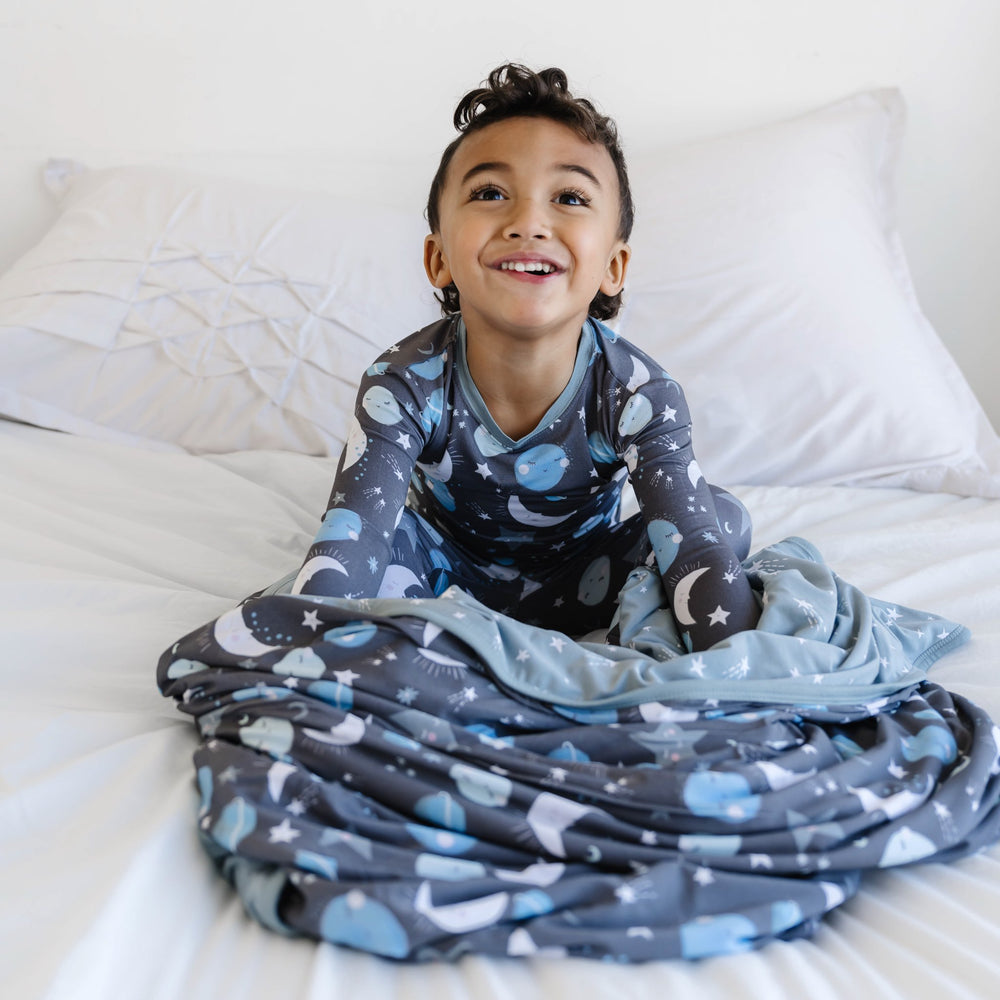 Image of little boy sitting with his large cloud blanket in blue to the moon and back print. This print features blue and gray moons, stars, and planets on a charcoal background with dusty gray trim.