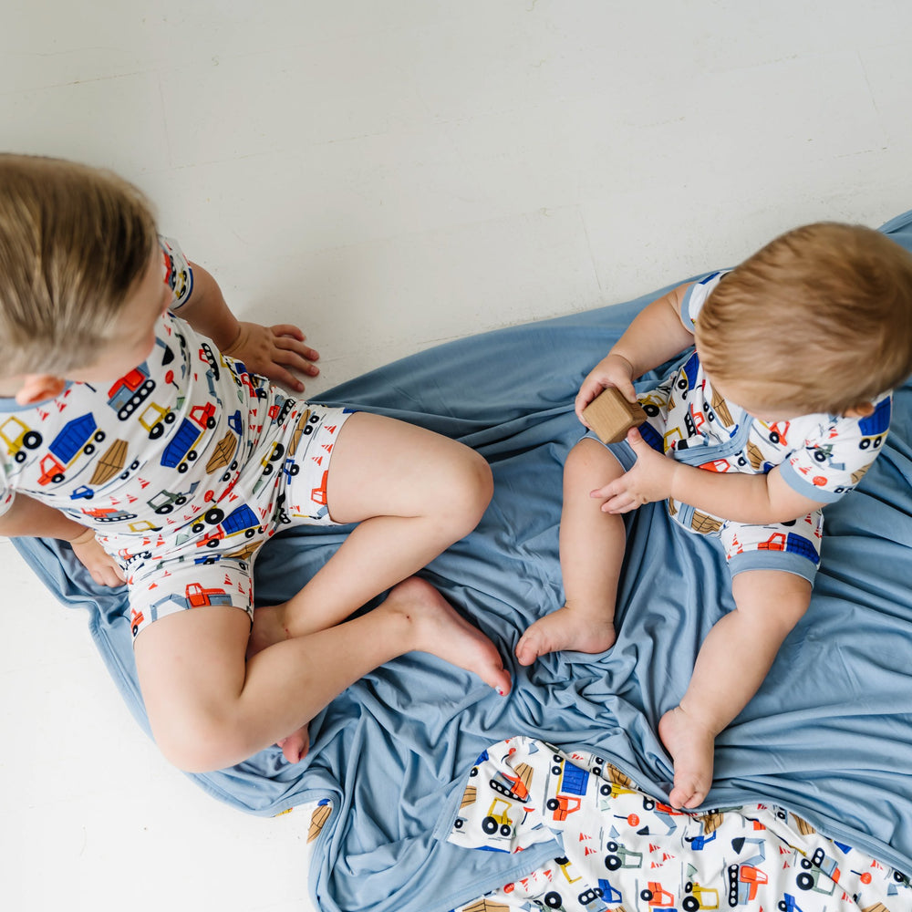 Image of two little boys wearing matching short sleeve and shorts zip romper and pajama sets in Construction print. They are playing with wooden blocks while sitting atop a matching construction blanket. The construction print is tractors and construction trucks atop a white background with light blue trim.