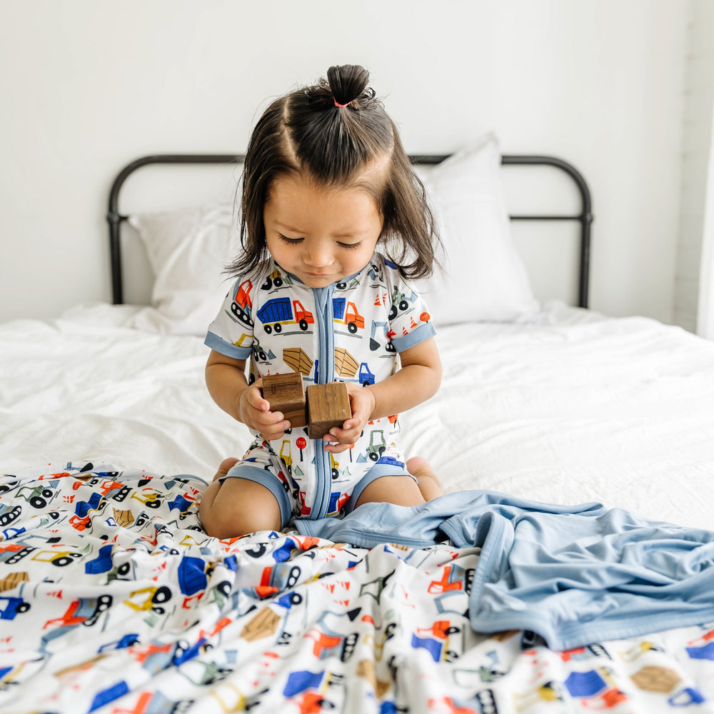 Image of little boy wearing short sleeve and shorts zip romper in Construction print and playing with wooden blocks on the bed while sitting atop his matching construction blanket. The construction print is tractors and construction trucks atop a white background with light blue trim.