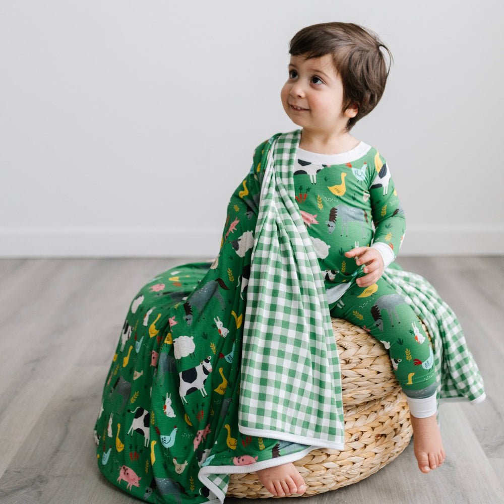 Click to see full screen - Image of toddler boy sitting on a rattan pouf with a green farm animals blanket draped over his shoulder. He is shown wearing a two piece pajama set in green farm animals print. The blanket features a double-sided mix and match print, with one side showin