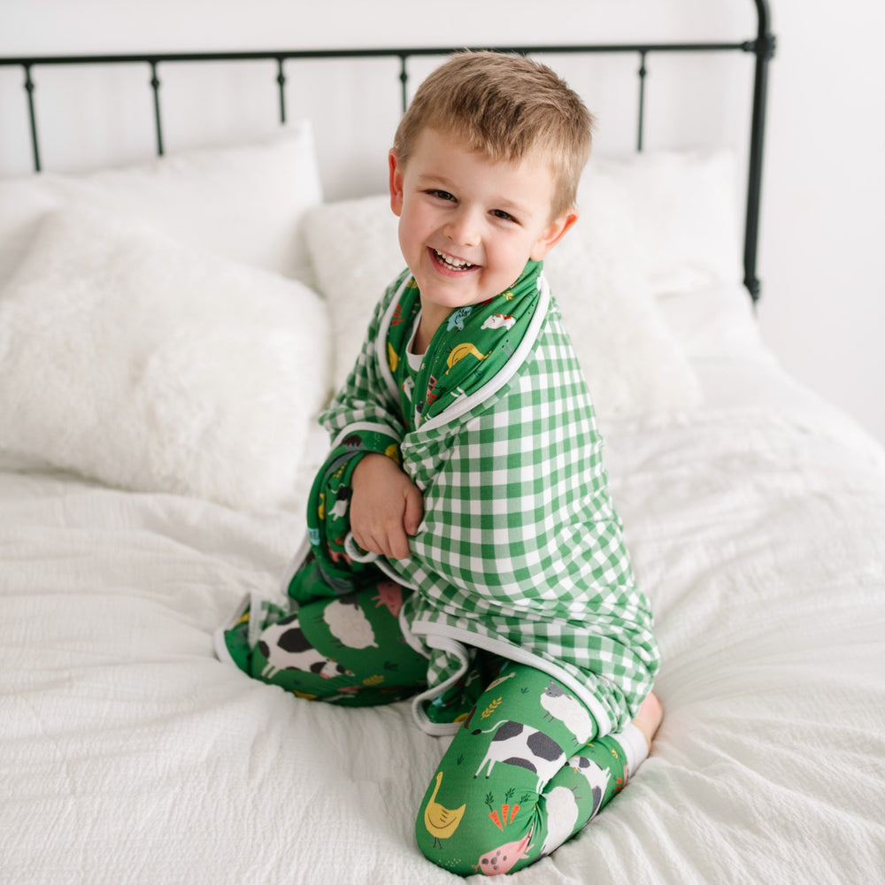 Image of little boy wrapping a farm animal printed blanket around him. He is shown wearing a two-piece pajama set in green farm animals print. The blanket features a double-sided mix and match print, with one side showing the green farm animals and the ot