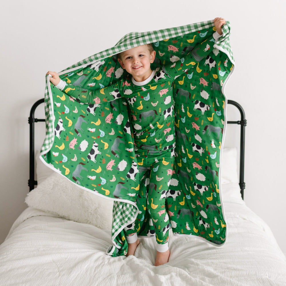 Image of little boy standing on the bed holding a farm animal printed blanket over his head. He is shown wearing a two-piece pajama set, also in a green farm animals print. The blanket features a double-sided mix and match print, with one side showing the