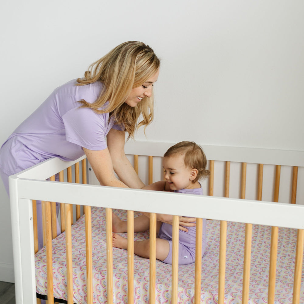 Image of mother bending down to grab her infant, both wearing matching Wisteria purple pajama sets. The infant baby is sitting in a white crib on top of a Pastel Rainbows fitted crib sheet. 