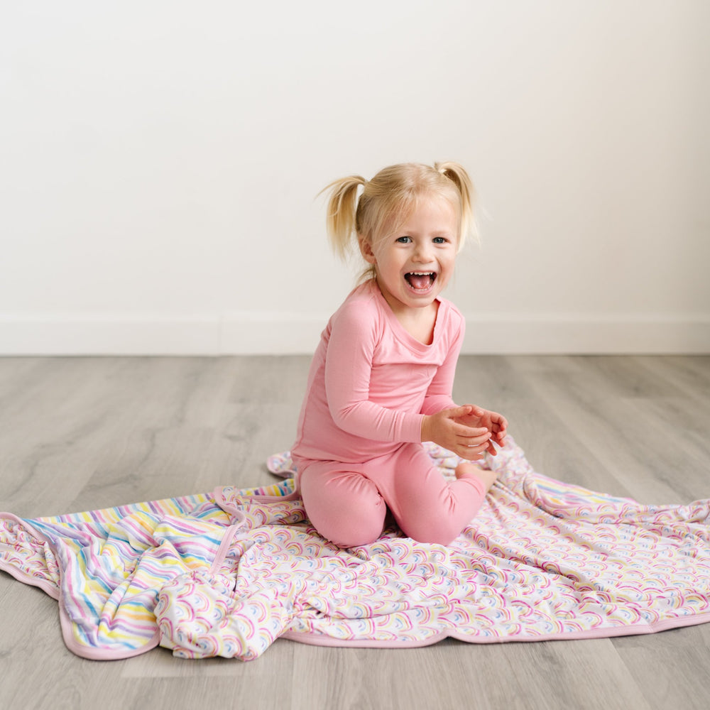 Child in pink two-piece pajama set sitting on double-sided rainbow and stripe printed blanket