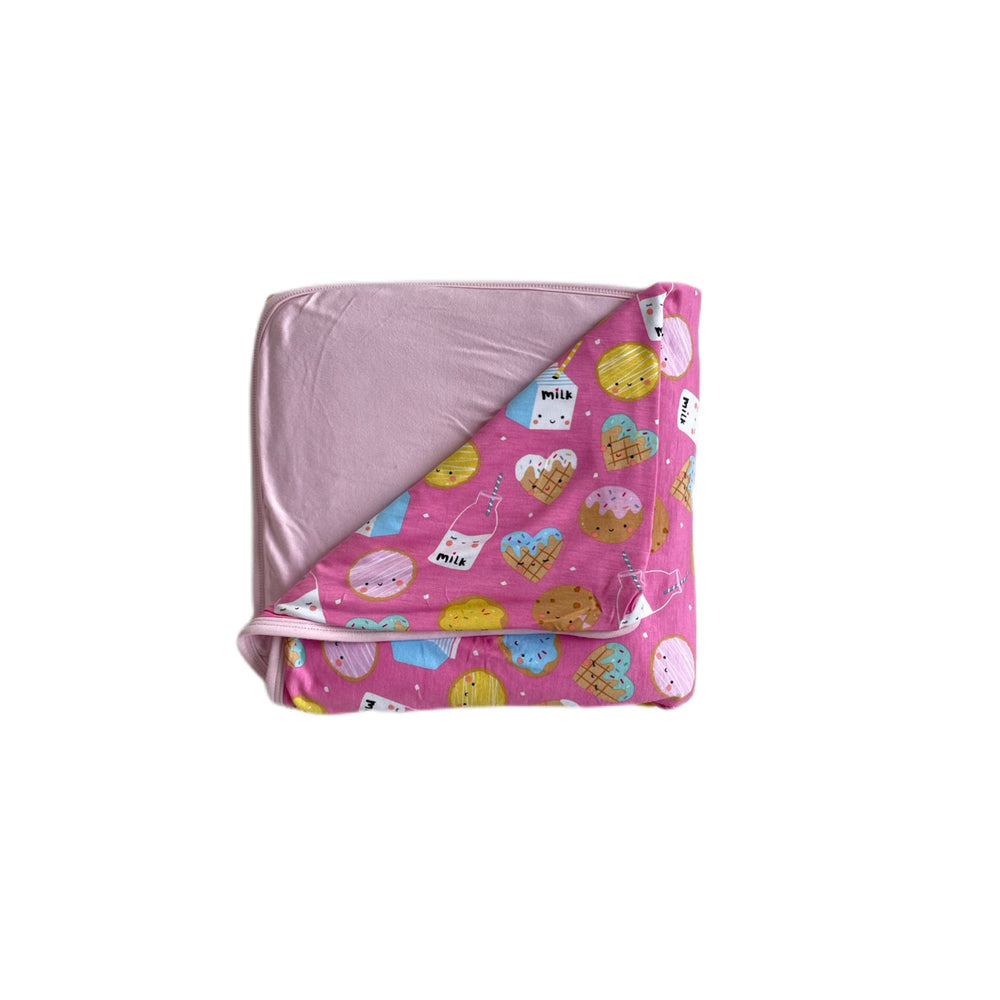 Flat lay image of triple layer large cloud blanket in Pink Cookies and Milk print. This blanket features a double-sided design, with one side that shows a variety of different cookies with bottles and boxes of milk on a pink background and the other side featuring a coordinating solid light pink.