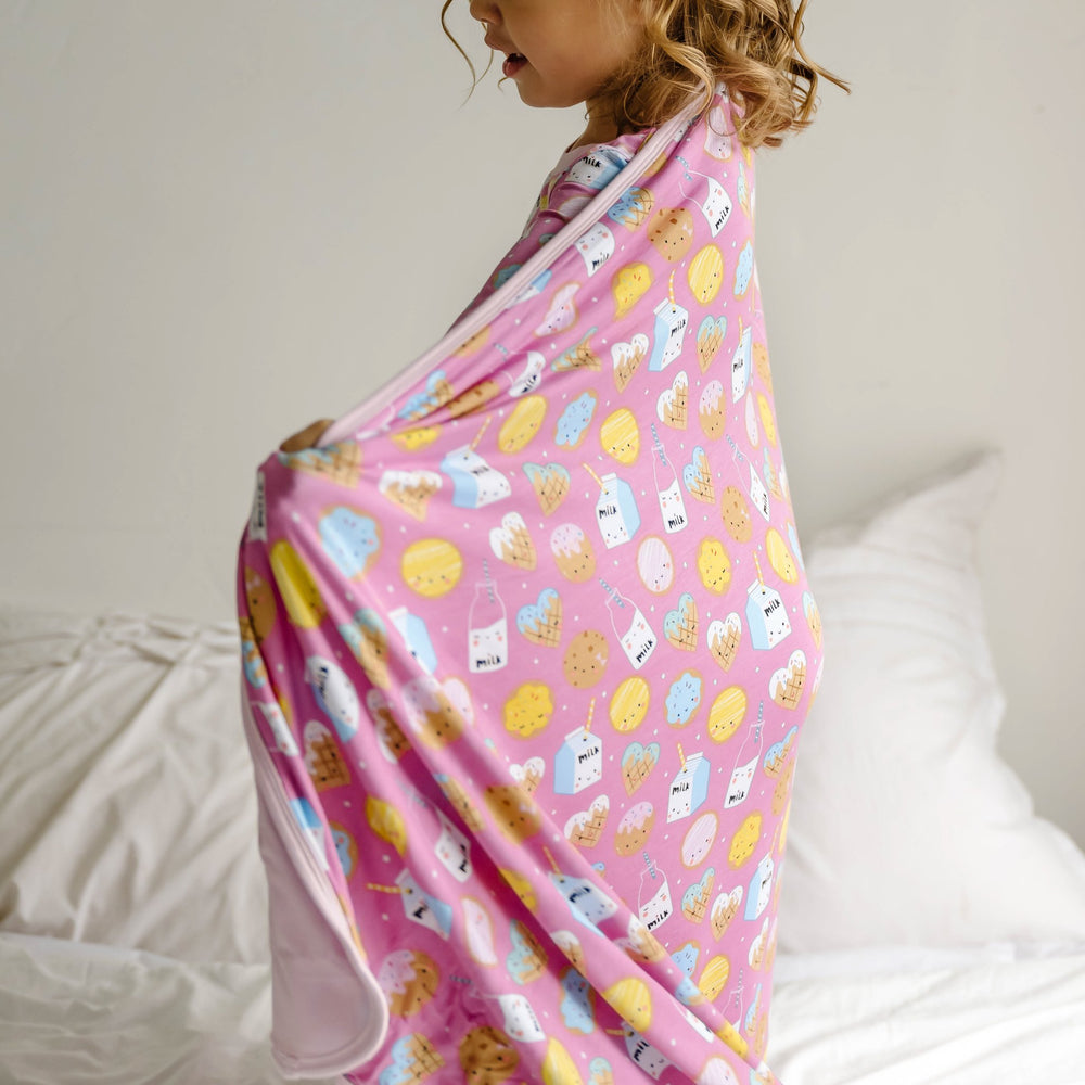 Image of little girl draping a Pink Cookies and Milk blanket around her. This blanket features a double-sided design, with one side that shows a variety of different cookies with bottles and boxes of milk on a pink background and the other side featuring a coordinating solid light pink.