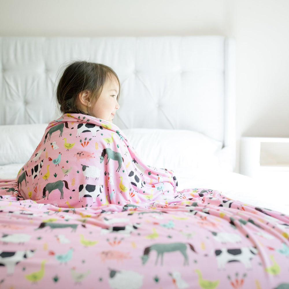 Image of little girl sitting on a bed with a farm animal printed blanket wrapped around her. She is shown wearing matching farm animal printed pajamas. The blanket features a double-sided mix and match print, with one side showing the pink farm animals an