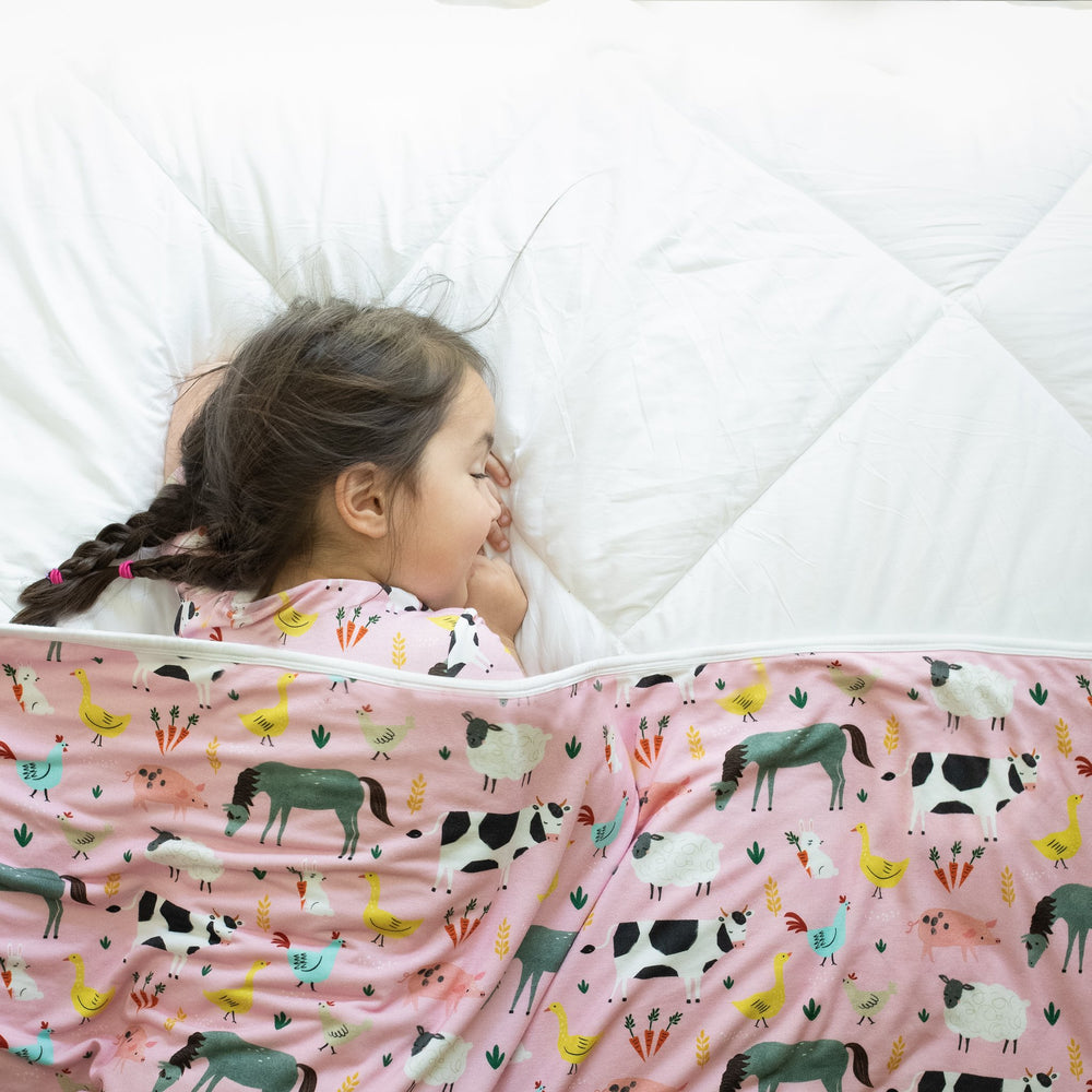 Image of little girl sleeping with a farm animal printed blanket draped over her. She is shown wearing matching farm animal printed pajamas. The blanket features a double-sided mix and match print, with one side showing the pink farm animals and the other
