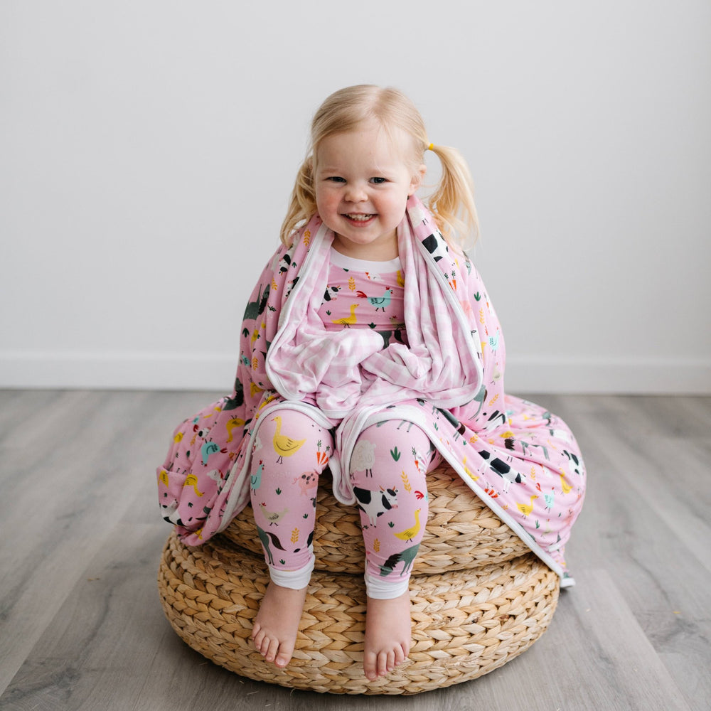 Image of little girl sitting on a stack of rattan poufs with a pink farm animal printed blanket wrapped around her. She is shown wearing matching farm animal printed pajamas. The blanket features a double-sided mix and match print, with one side showing t