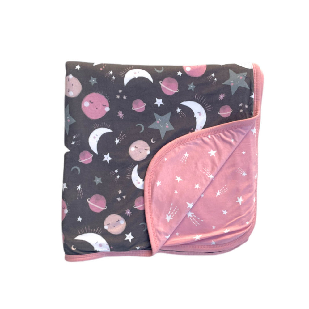 Flat lay image of large cloud blanket in pink to the moon and back print. This blanket features a double-sided design, with pink and gray moons, stars, and planets on a charcoal background on one side and white shooting stars on a coordinating pink backgr