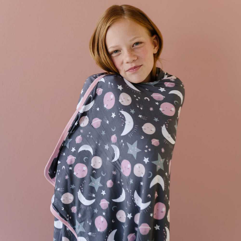 Image of little girl holding her large cloud blanket in pink to the moon and back print. This blanket features a double-sided design, with pink and gray moons, stars, and planets on a charcoal background on one side and white shooting stars on a coordinat