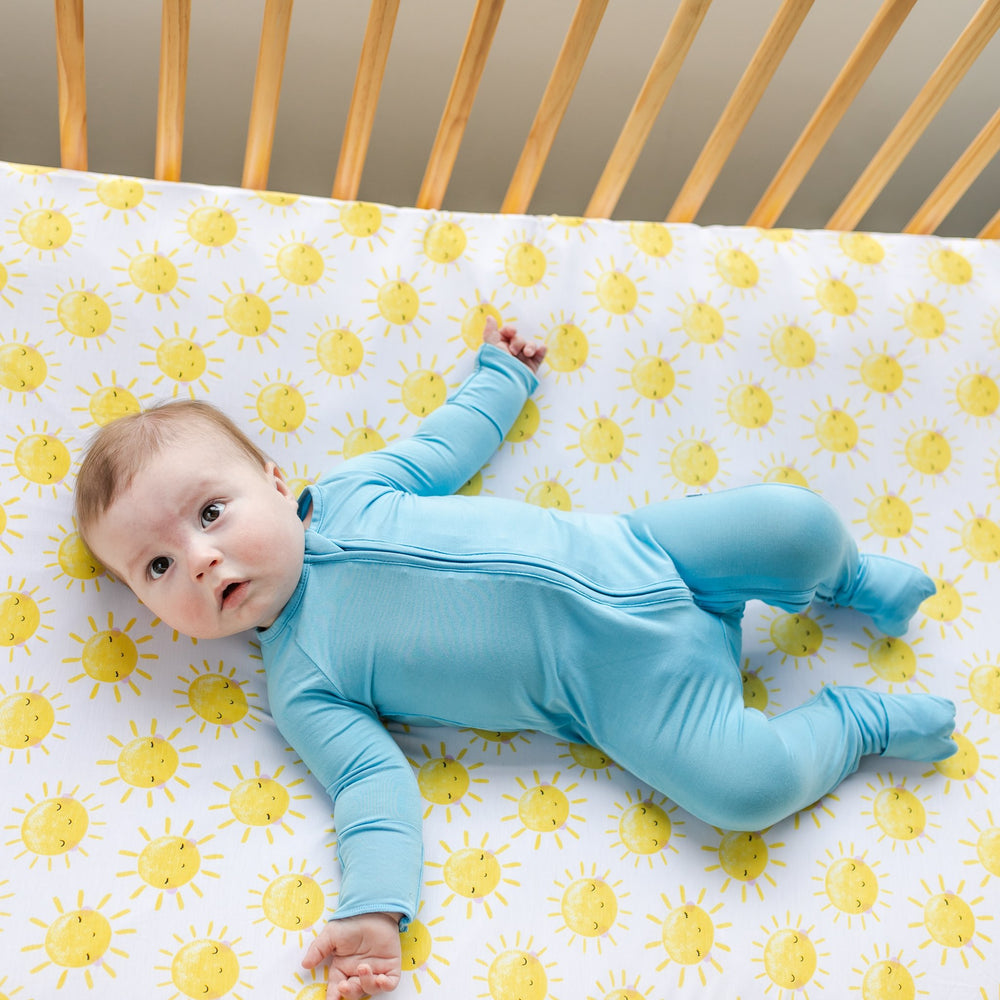 Image of infant wearing a sky blue zip romper laying on a yellow sunshine printed crib sheet