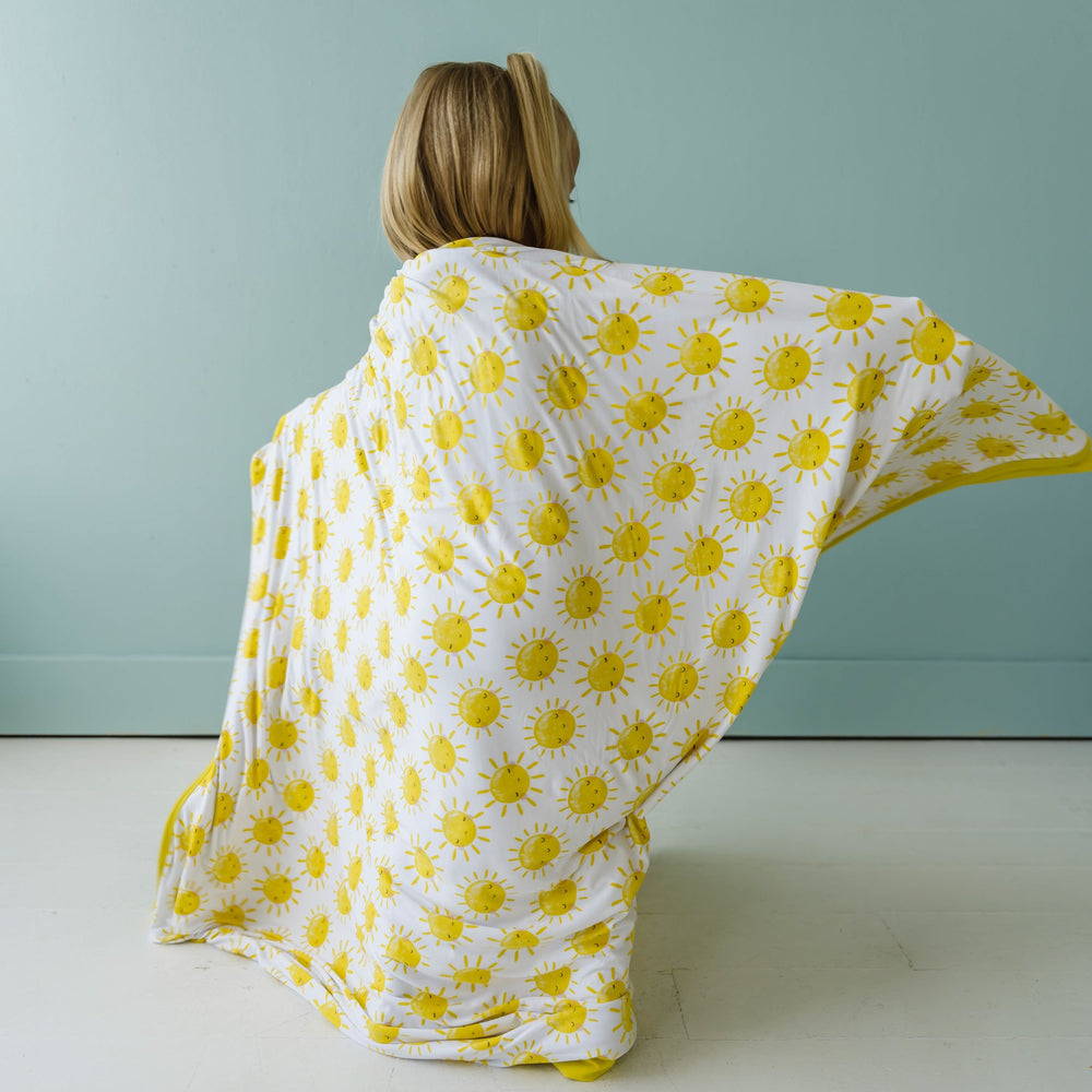Image of little girl draping a sunshine printed, triple-layered blanket over herself. This print features yellow smiling suns that sit on a white background with yellow trim.