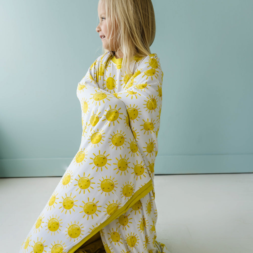 Image of little girl draping a sunshine printed, triple-layered blanket over herself. This print features yellow smiling suns that sit on a white background with yellow trim.
