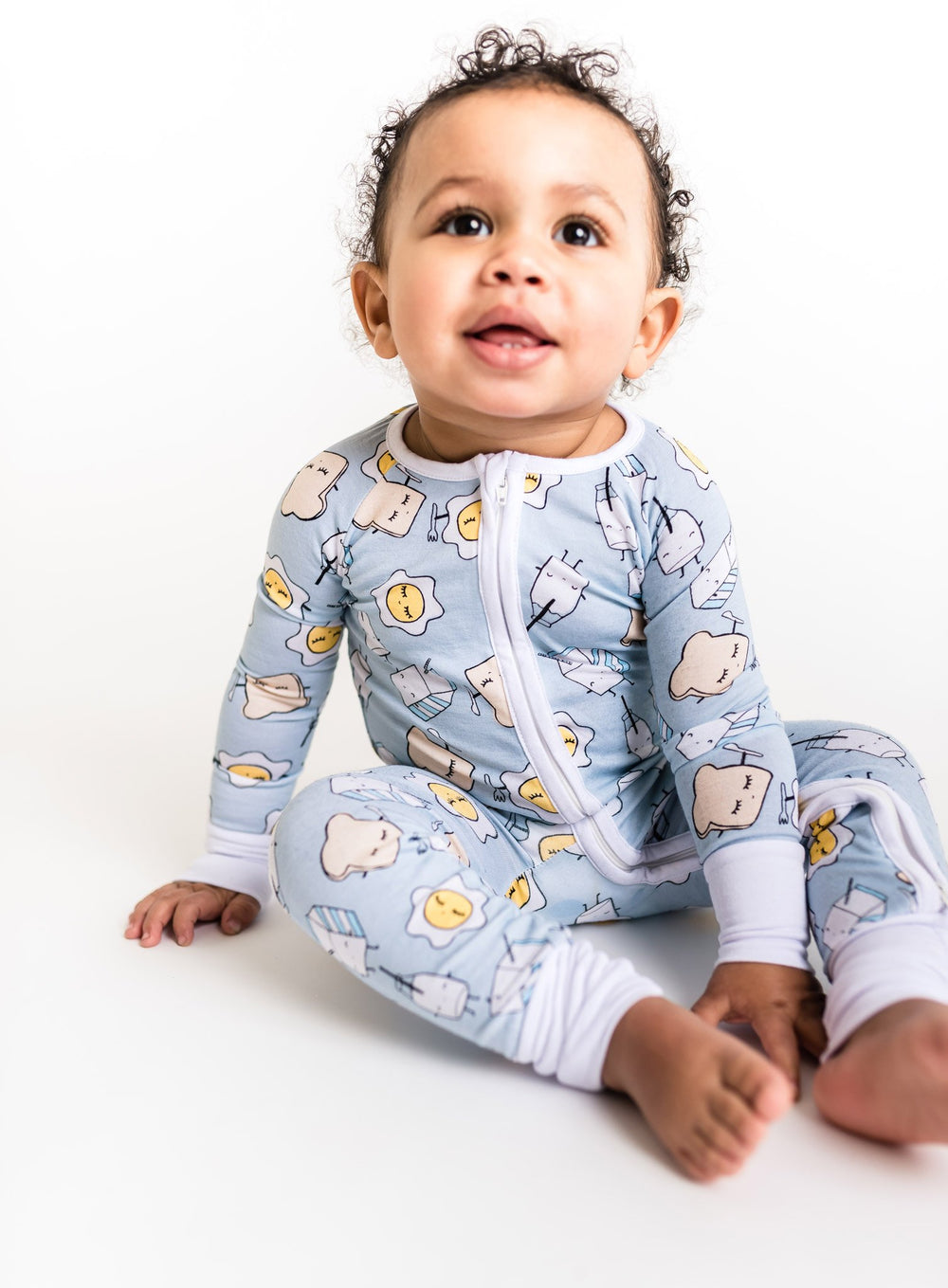 Image of baby boy wearing Blue Breakfast Buddies printed Zippy. This print has a light blue background with white trim accents and the breakfast foods featured on this print include sunny side up eggs, toast, and milk.