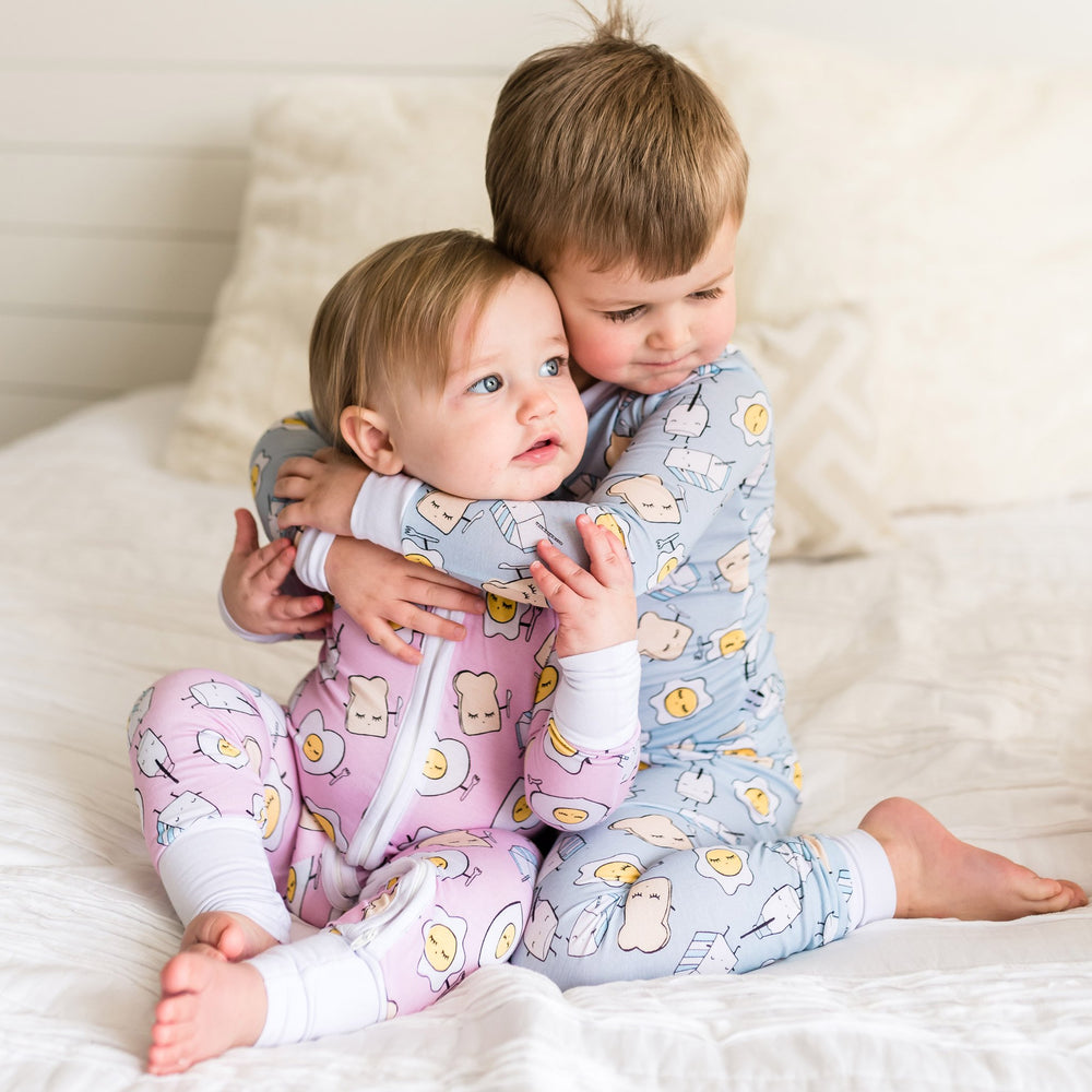 Image of brother hugging his little sister. Both are wearing matching Breakfast Buddies printed pajamas. Toddler boy is wearing Blue Breakfast Buddies printed pajamas with white trim accents, while little girl is wearing Pink Breakfast Buddies printed Zippy. The breakfast foods featured on this print include sunny side up eggs, toast, and milk.