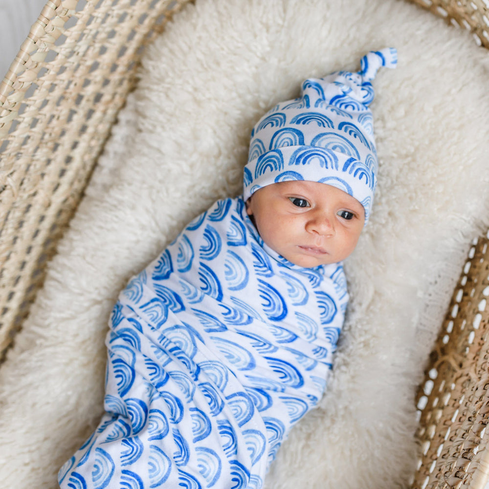 Image of newborn baby boy in rainbow printed swaddle and hat set. This print sits on a white background with shades of blue rainbows and sky blue trim details.
