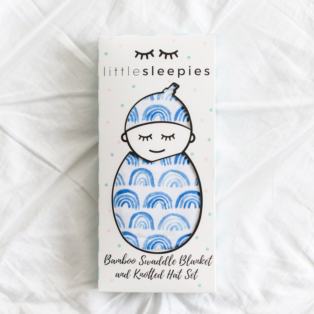 Image of blue rainbow printed swaddle and hat set in Little Sleepies packaging. This print sits on a white background with shades of blue rainbows and sky blue trim details.