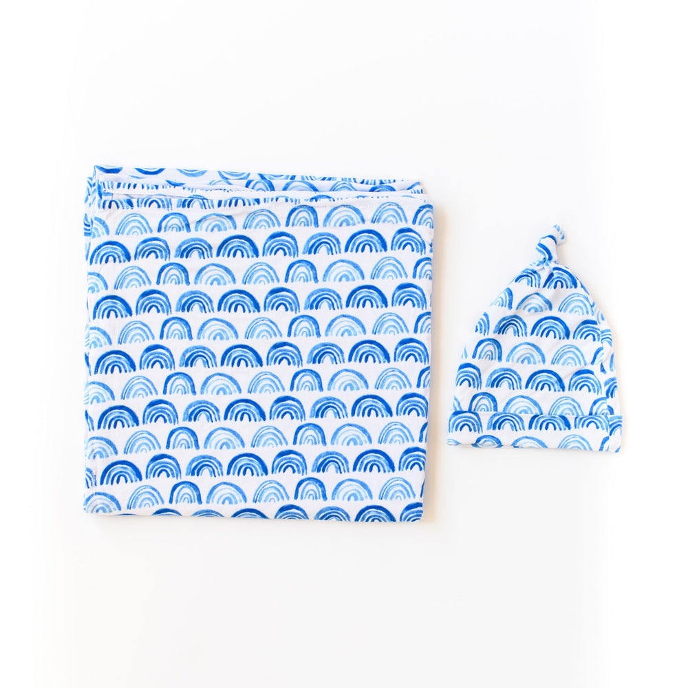 Flat lay photo of blue rainbow printed swaddle and hat set. This print sits on a white background with shades of blue rainbows and sky blue trim details.