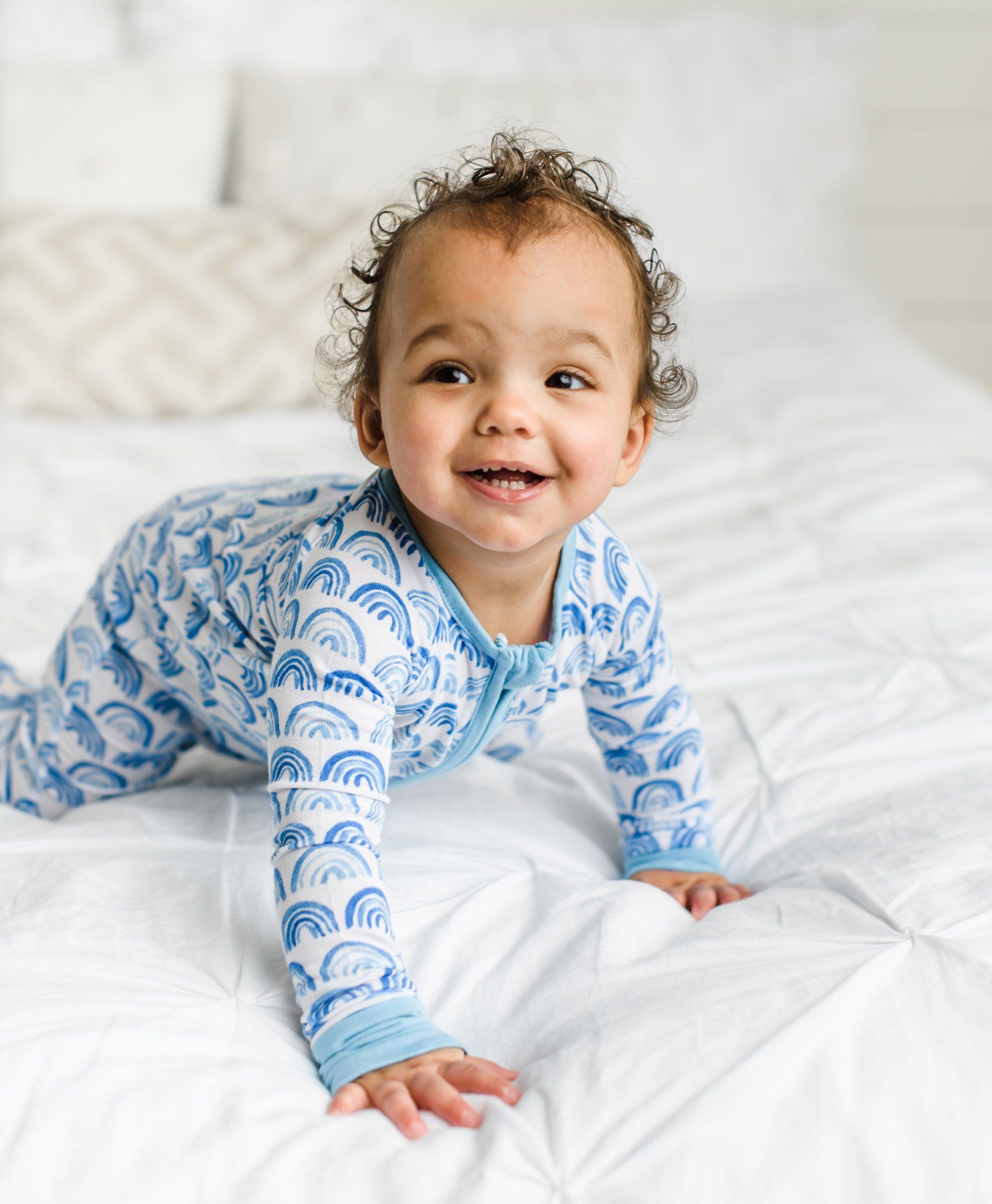 Image of baby boy wearing blue rainbow printed zip up romper. This print sits on a white background with shades of blue rainbows and sky blue trim details.