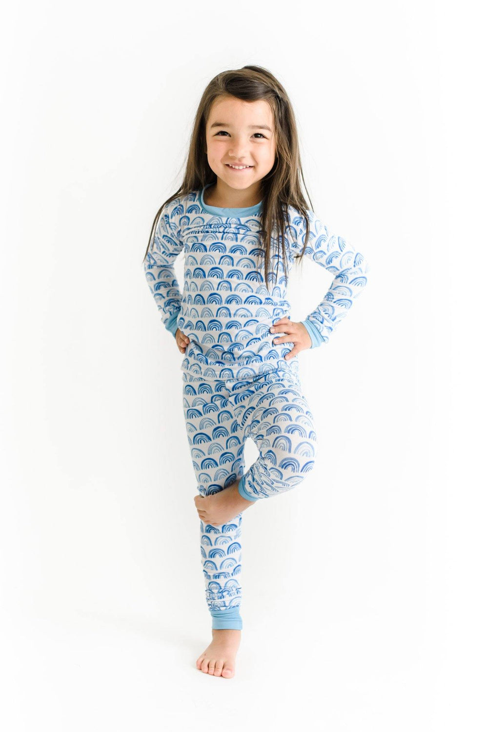 Image of toddler girl wearing rainbow printed pajama set. This print sits on a white background with shades of blue rainbows and sky blue trim details.