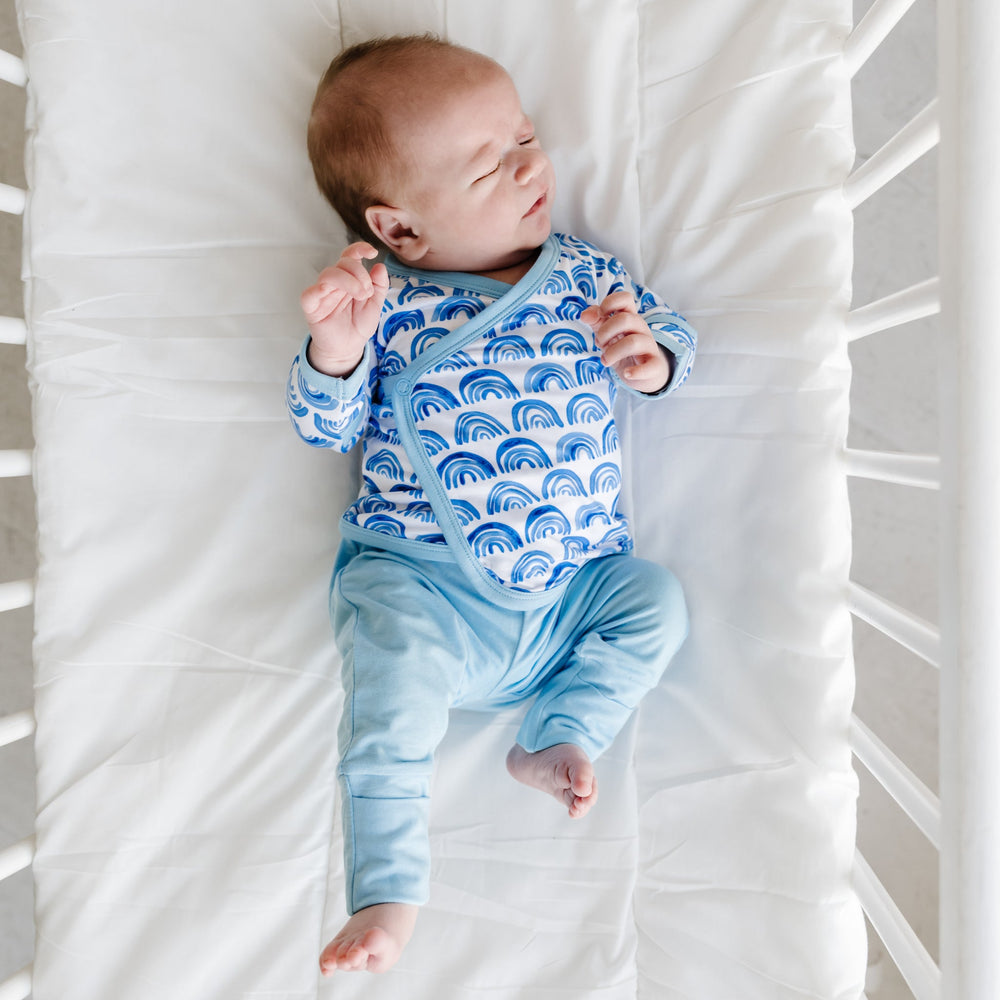 Image of an infant wearing a two-piece crossover set that features a long sleeve wrap style top with snap closures and coordinating pants with convertible footies. This photo features the wrap style top in Blue Rainbows print that features watercolor style rainbows in a bright blue on a white background with soft blue trim.