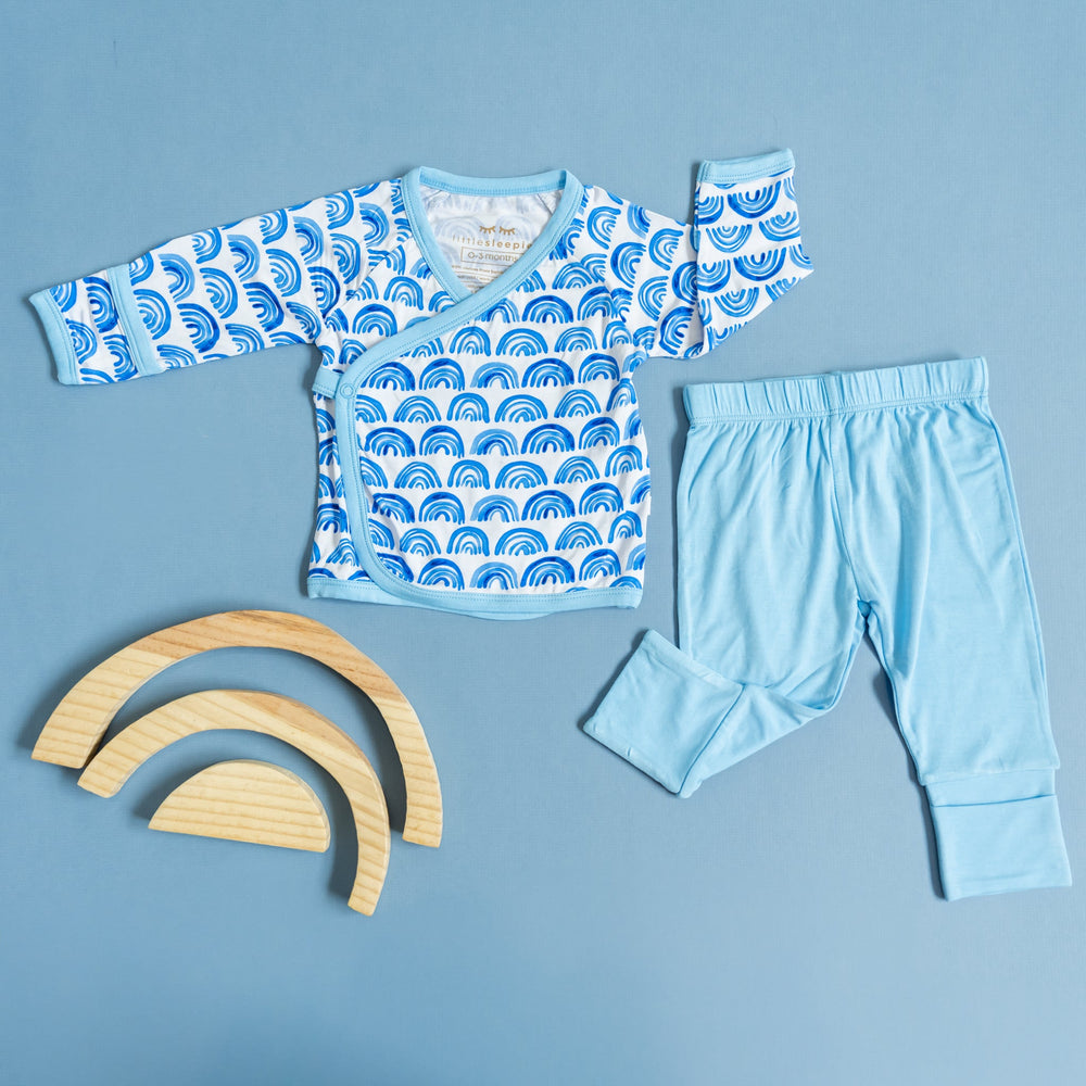 Flat lay image of a two-piece crossover set that features a long sleeve wrap style top with snap closures and coordinating pants with convertible footies. This style pictured has the wrap style top in Blue Rainbows print with watercolor style rainbows in a bright blue on a white background with soft blue trim.