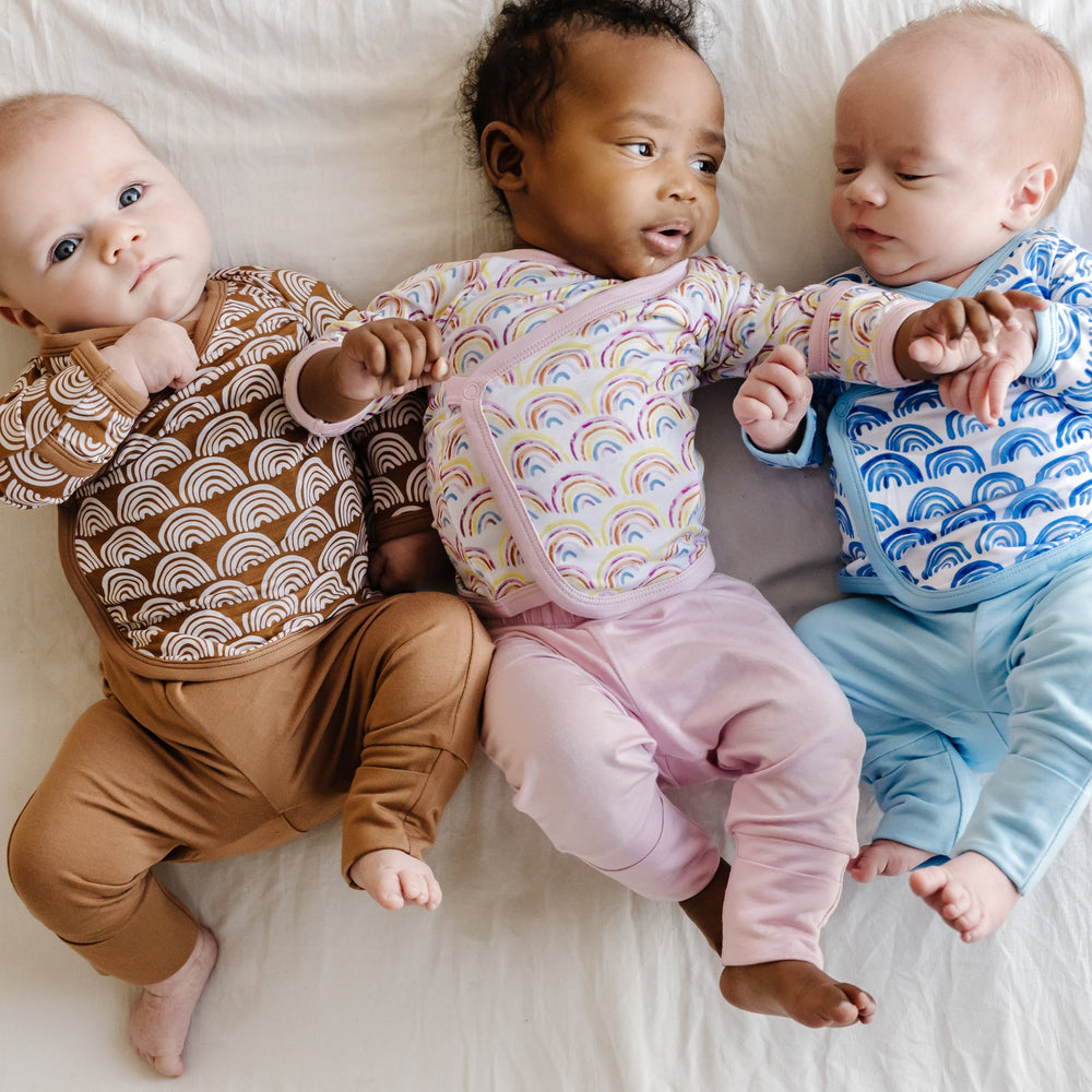 Image of three infants wearing two-piece crossover sets that features a long sleeve wrap style top with snap closures and coordinating pants with convertible footies. This photo features wrap style tops from the Rainbows collection with Rust Rainbows, Pastel Rainbows, and Blue Rainbows print.