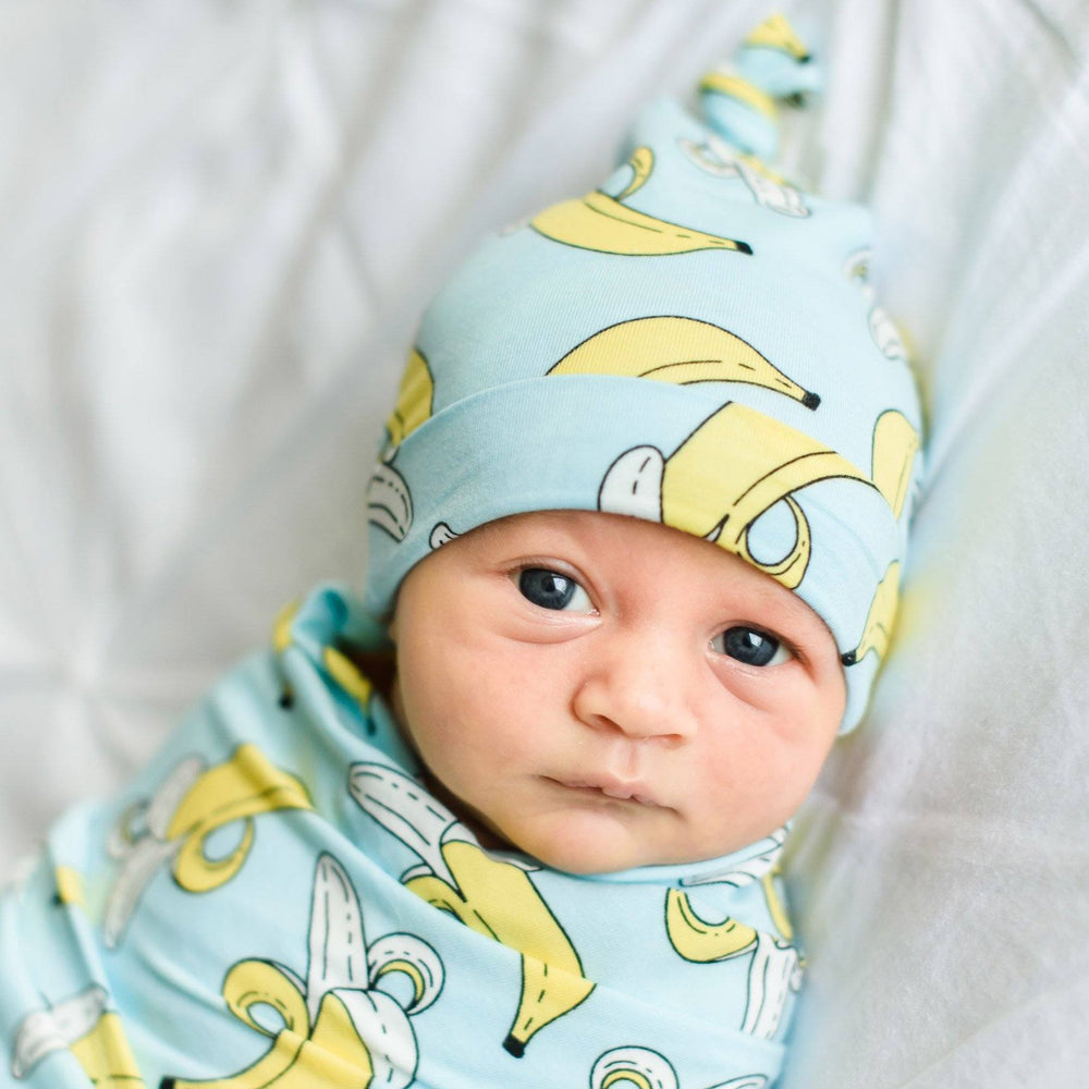 Baby boy wrapped up in banana printed swaddle. He is also wearing the matching banana printed hat. This set features a light blue background with pops of yellow coming from the bananas.