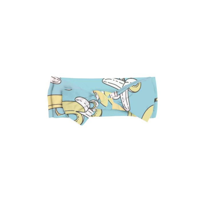 Flat lay photo of banana printed bow headband. The bow headband has a light blue background with pops of yellow coming from the banana print.