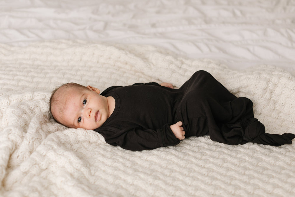 Baby boy lying down and wearing solid black knotted gown. Image shows the gown tied at the bottom to cover the baby's feet.