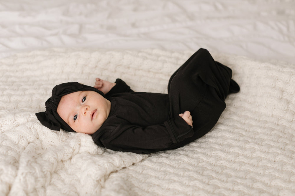 Baby girl lying down and wearing solid black knotted gown and matching solid black bow headband. Image shows the gown tied at the bottom to cover the baby's feet.