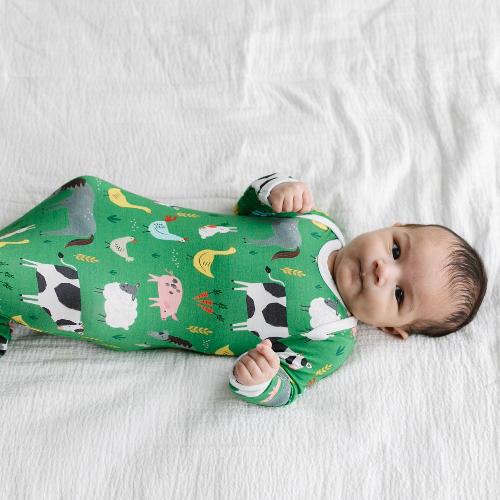 Image of infant boy wearing a knotted gown in green farm animals print. This print includes a green background with white trim details. The farm animals featured on this print include cows, pigs, ducks, sheep, pigs, chickens, and bunnies. 