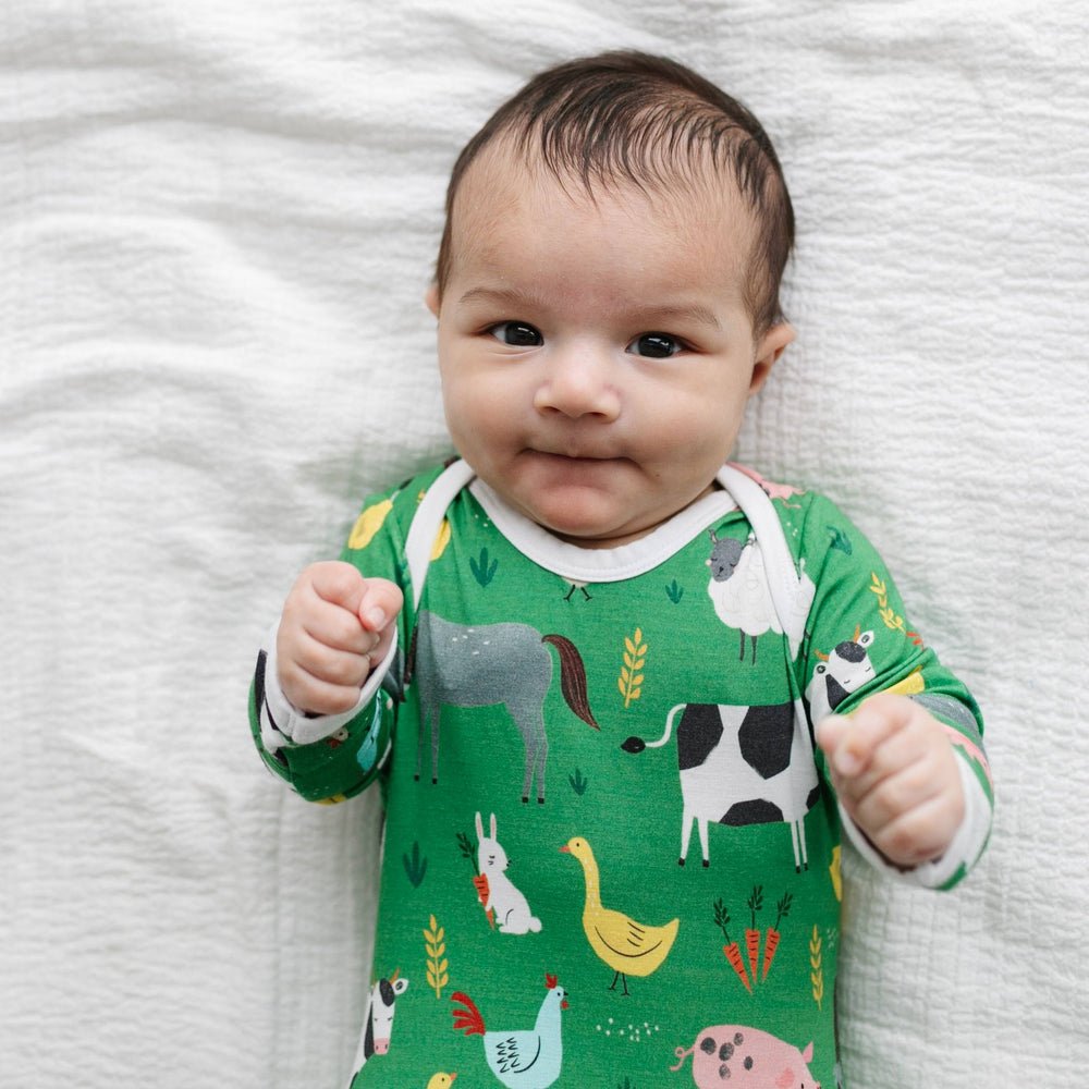 Image of infant boy wearing a knotted gown in green farm animals print. This print includes a green background with white trim details. The farm animals featured on this print include cows, pigs, ducks, sheep, pigs, chickens, and bunnies.