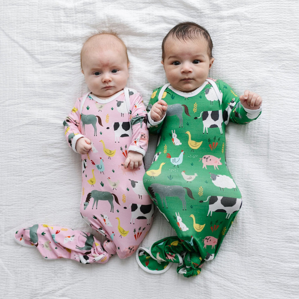 Image of infant boy and girl wearing matching farm animal printed knotted gowns. The infant boy is seen wearing a green gown, and the infant girl is shown wearing pink. The farm animals featured on this print include cows, pigs, ducks, sheep, pigs, chicke