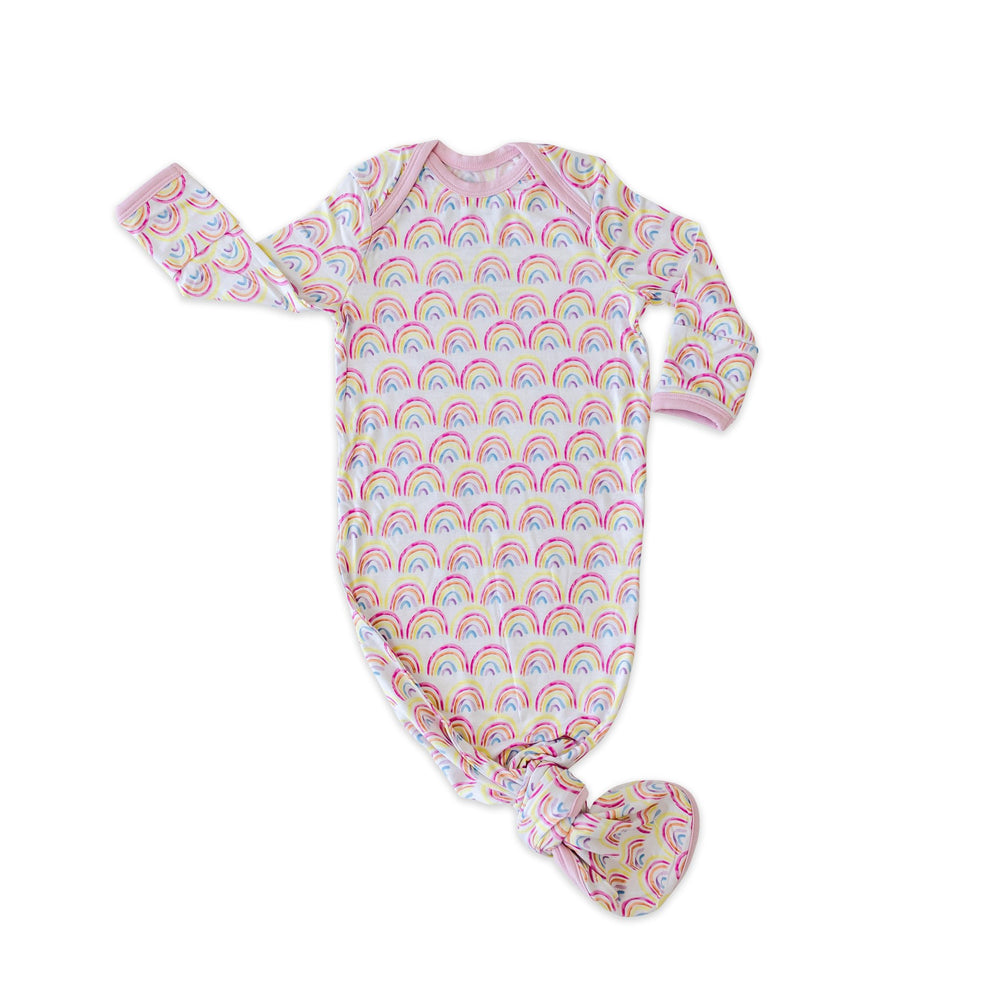 Flat lay image of infant knotted gown in rainbow print.  This print features multicolored rainbows that sit upon a white background with light pink accent trim.