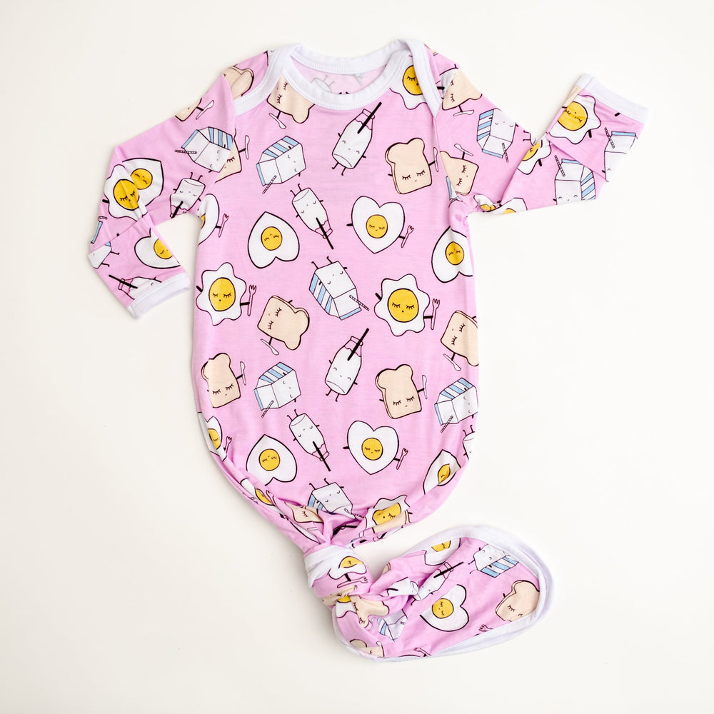 Flat lay image of infant knotted gown in Pink Breakfast Buddies print. This print has a light pink background with white trim accents and the breakfast foods featured on this print include sunny side up eggs, toast, and milk.