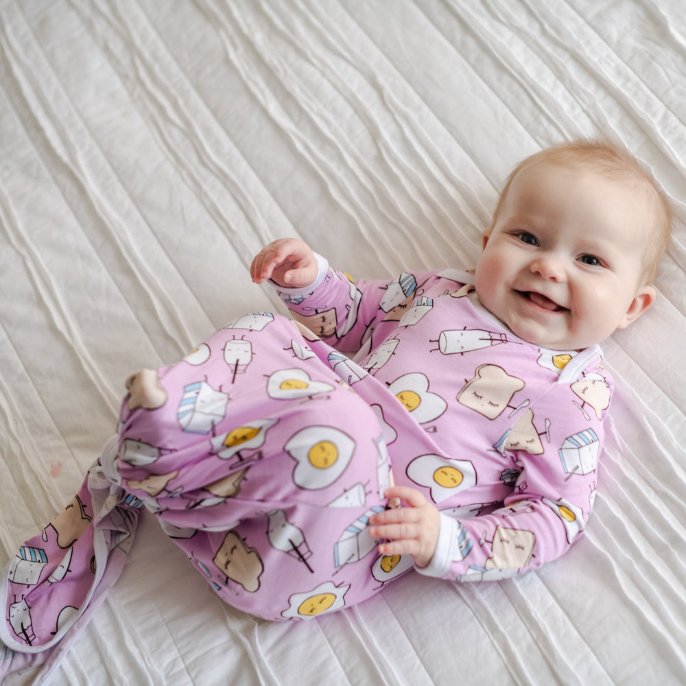 Image of infant girl wearing knotted gown in Pink Breakfast Buddies print. This print has a light pink background with white trim accents and the breakfast foods featured on this print include sunny side up eggs, toast, and milk.