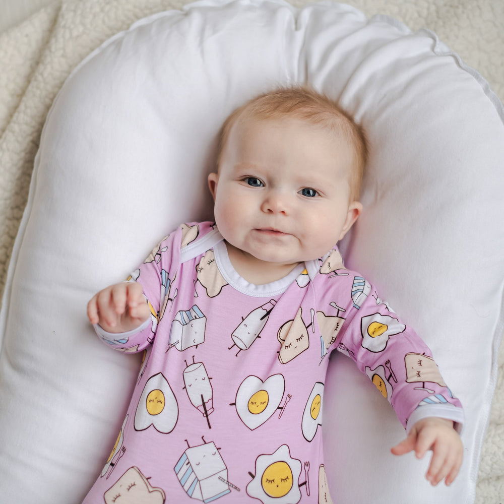 Image of infant girl wearing knotted gown in Pink Breakfast Buddies print. This print has a light pink background with white trim accents and the breakfast foods featured on this print include sunny side up eggs, toast, and milk.