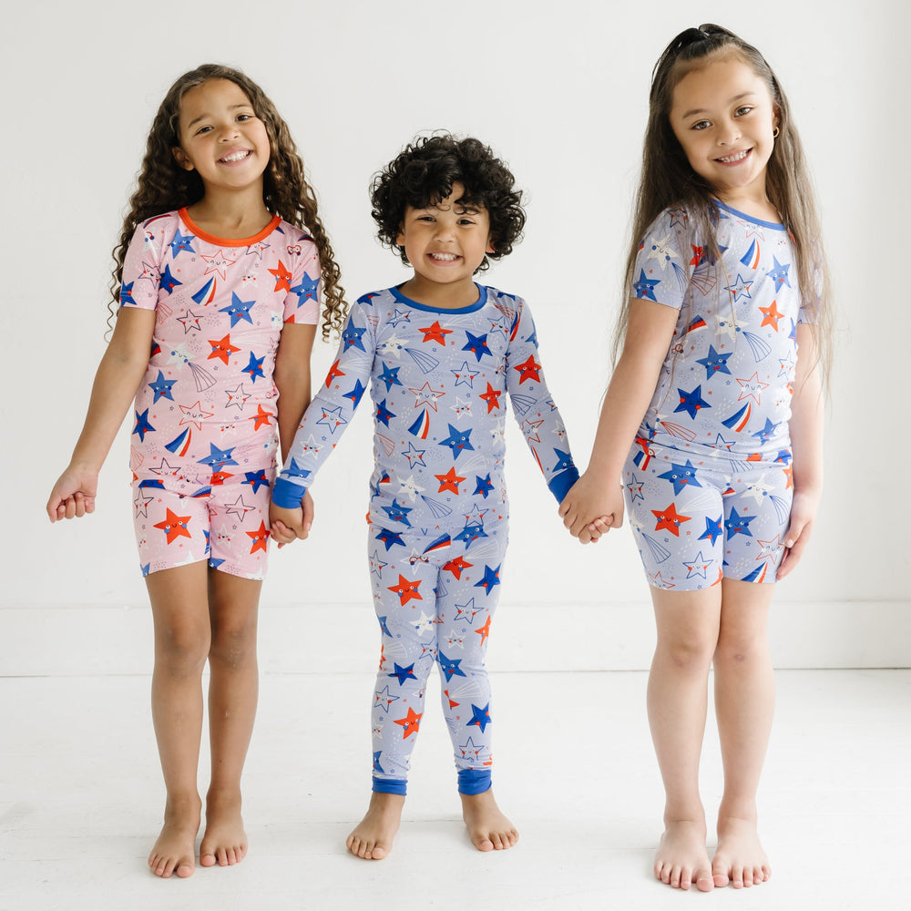 Three children holding hands wearing coordinating Stars and Stripes pajamas. Two children are wearing Pink and Blue Stars and Stripes printed short sleeve and shorts pajama sets and one child is wearing a Blue Stars and Stripes printed two piece pajama set 