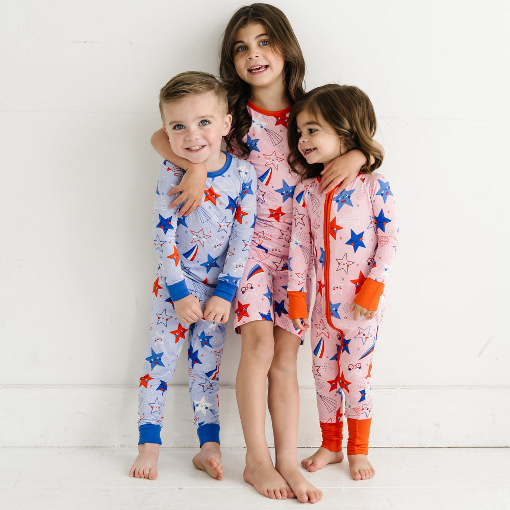 Three children hugging. One child is wearing a Blue Stars and Stripes printed two piece pajama set and two children are wearing Pink Stars and Stripes printed two piece short sleeve and shorts pajama set and zippy