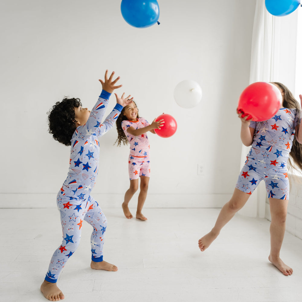 Three children playing together wearing coordinating Stars and Stripes pajamas. Two children are wearing Pink and Blue Stars and Stripes printed short sleeve and shorts pajama sets and one child is wearing a Blue Stars and Stripes printed two piece pajama set