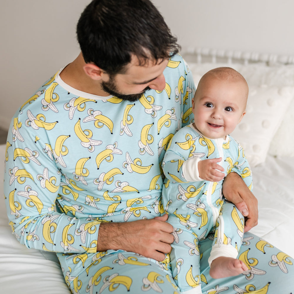 Father and son wearing matching banana printed pajamas. This print has a light blue background with pops of yellow coming from the bananas and also features white trim accents.