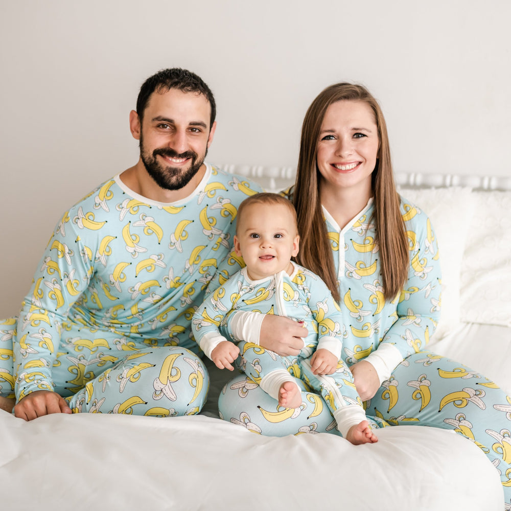 Family of three all wearing matching banana printed pajamas. The mom and dad pajama sets have a light blue background with white trim accents and pops of yellow coming from the banana print. While the baby boy's zip up romper has a light blue background with white trim accents and pops of yellow coming from the banana print